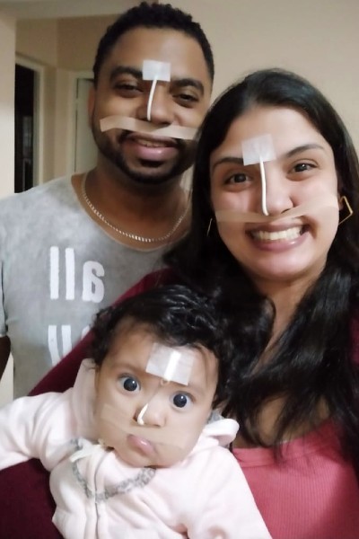 Foto da família. Gislaine, o marido e a bebê Melissa olham para a frente e esboçam um sorriso. A bebê usa um modelador nasal e os pais, em apoio à filha, colaram fita adesiva no rosto para imitar o aparelho